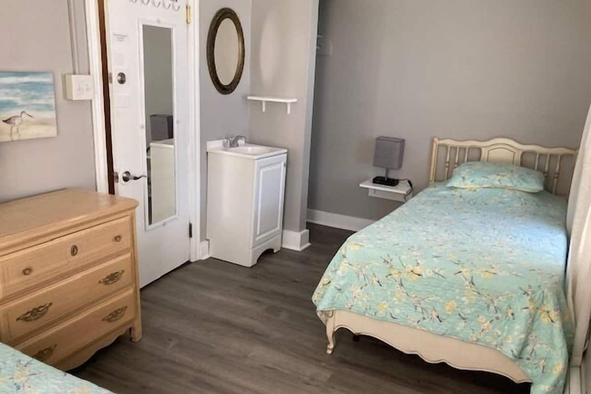 A bedroom with a wooden dresser, a bed with floral bedding, a nightstand with a lamp, and a mirror on the wall.