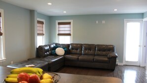 A living room with a sectional sofa, a dining table with a fruit bowl, and a door. - Ocean Park Inn (Ocean Grove)