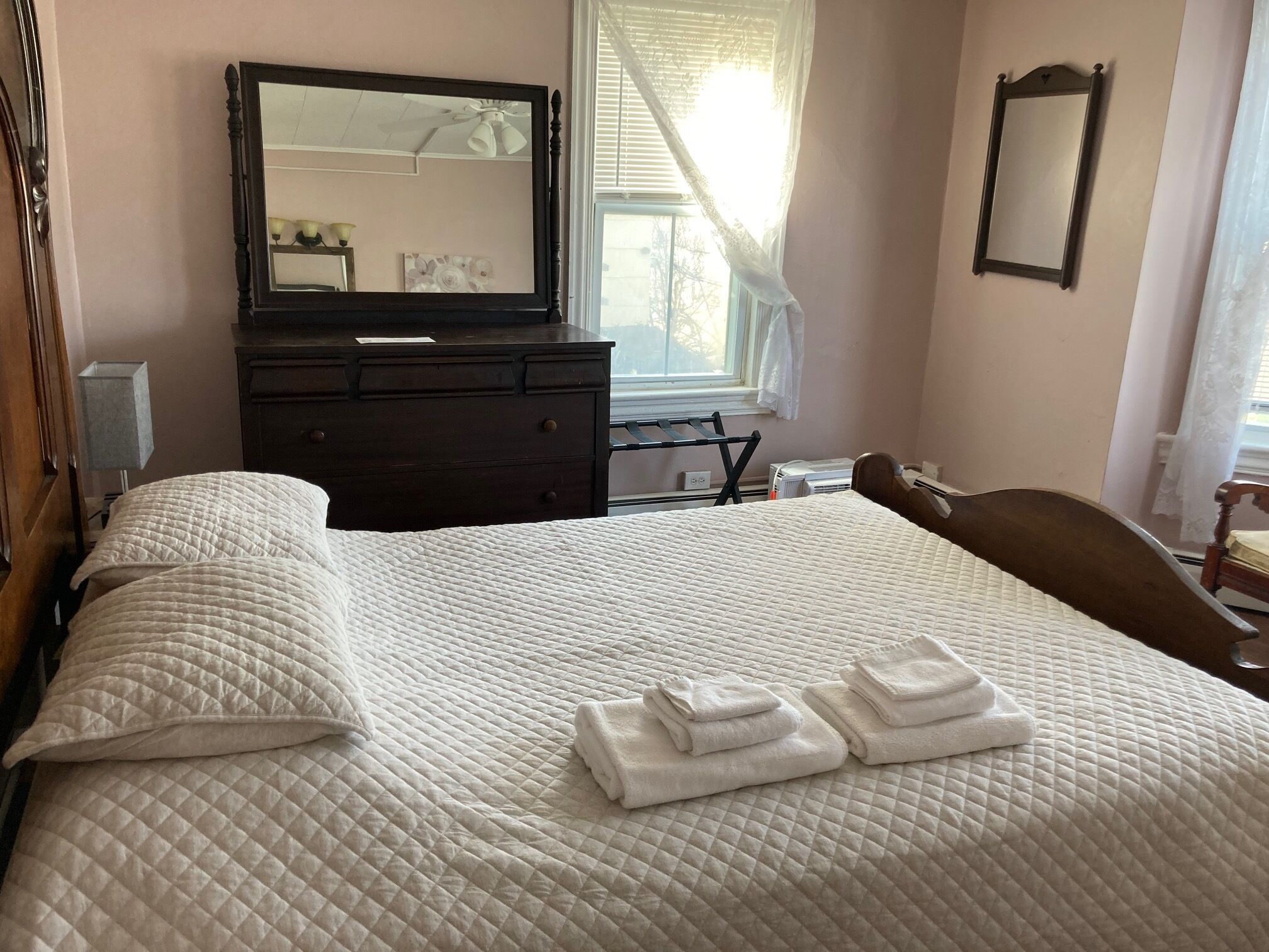 A neatly made bed with a quilted white comforter and two pillows, a dark wooden dresser with a mirror above it, and a window with sheer curtains allowing natural light into the room.