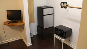 A compact room with a flat-screen TV on a wooden shelf, a small refrigerator, a microwave, and a broom leaning against the wall. - Summer Leaf (Reseda)