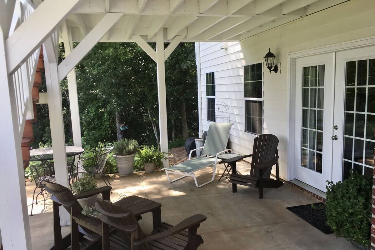 A covered patio with wooden furniture, potted plants, and a white house in the background.