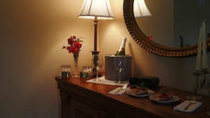 A wooden table with a lamp, a mirror, a vase with flowers, a bottle of champagne, and plates with food. - A Mighty Oak B&B (Pinnacle)