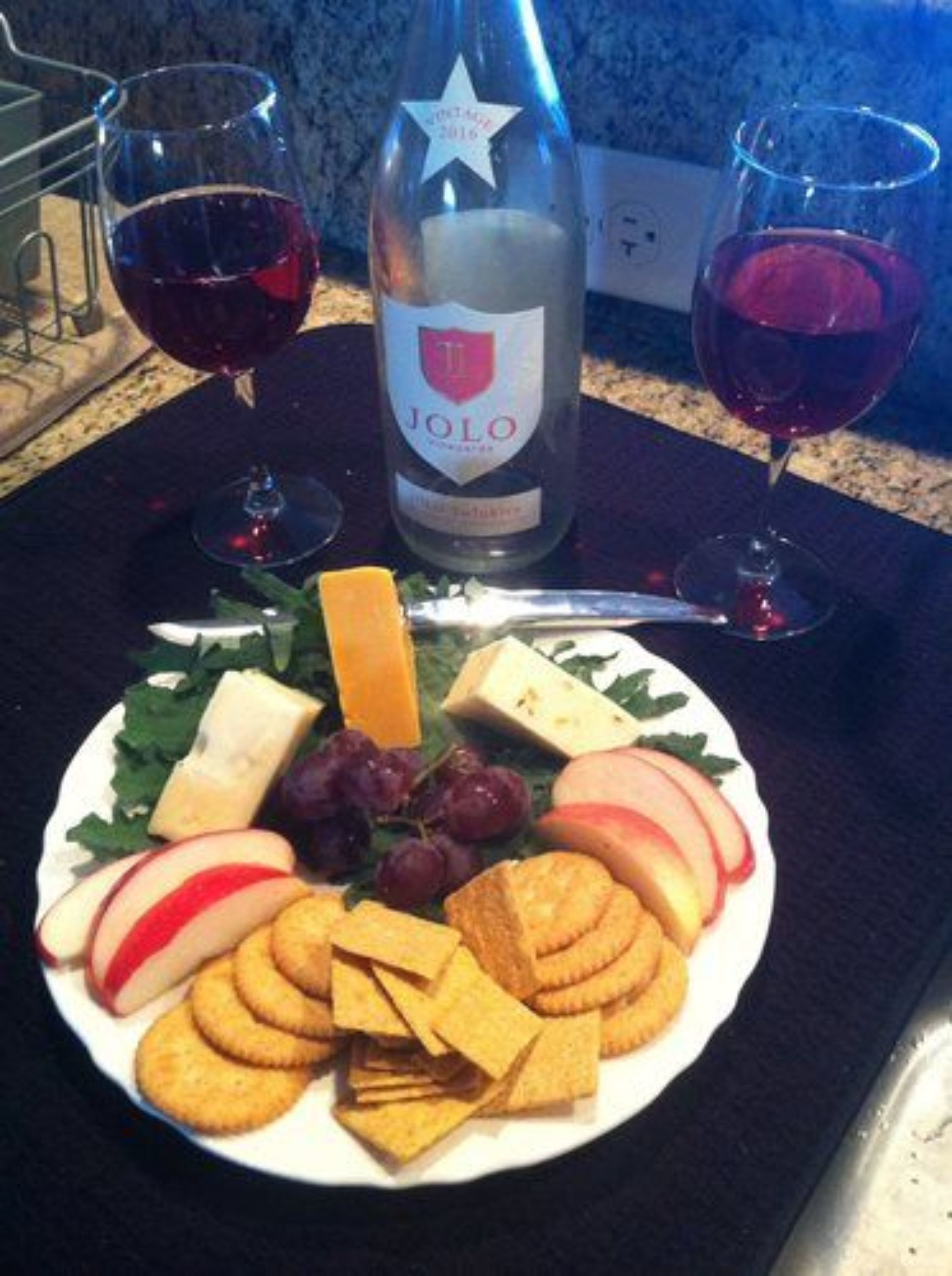 Un plato con queso, uvas, manzanas, galletas y una botella de vino Jolo.