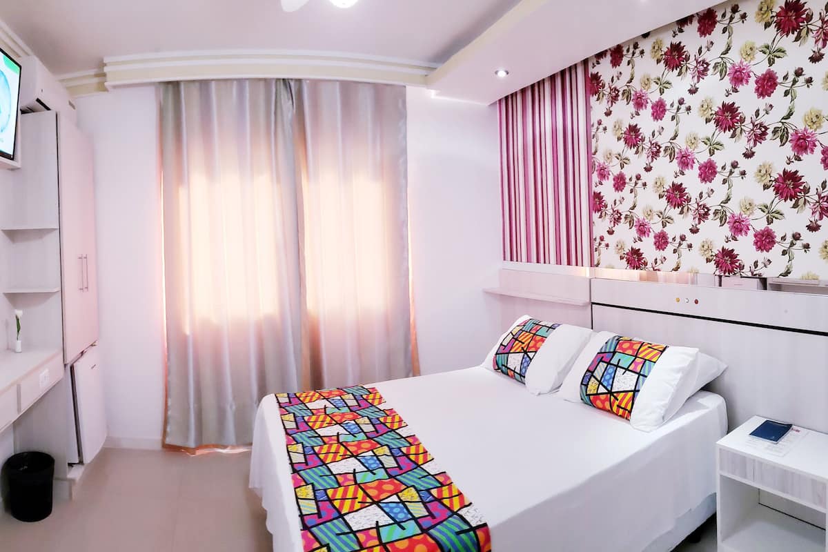 Ein Hotelzimmer mit einem Bett, einem an der Wand befestigten Fernseher, einem Spiegel, einem Fenster mit Vorhängen und floralem Wandpapier.