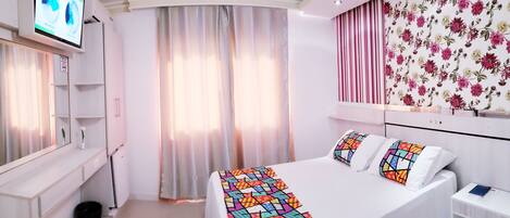 Habitación de hotel con cama, televisor montado en la pared, espejo, ventana con cortinas y un papel tapiz con motivos florales.
