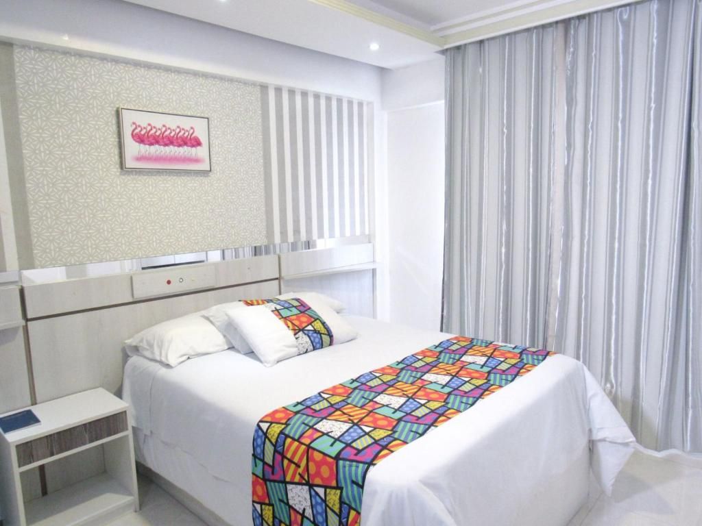 A hotel room with a bed, a nightstand, a wall-mounted shelf, and a window with curtains.