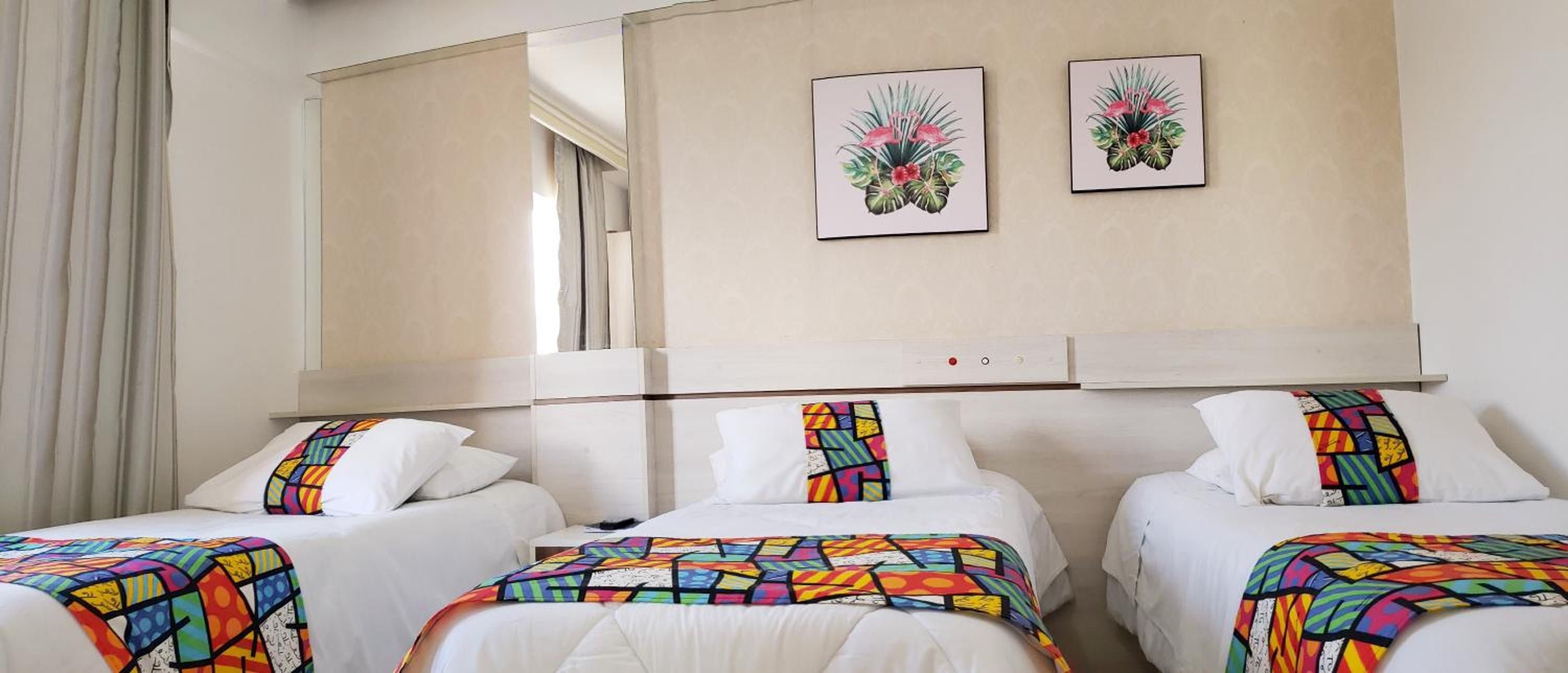 A hotel room with three beds, each with a colorful geometric-patterned bedspread. There are two framed floral paintings on the wall above the beds.