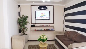 A modern living room with a sofa, armchair, and a coffee table with a vase of flowers. A flat-screen TV is mounted on the wall, displaying the hotel name. - Capão da Canoa - Hotel Rediadri (Capao da Canoa)