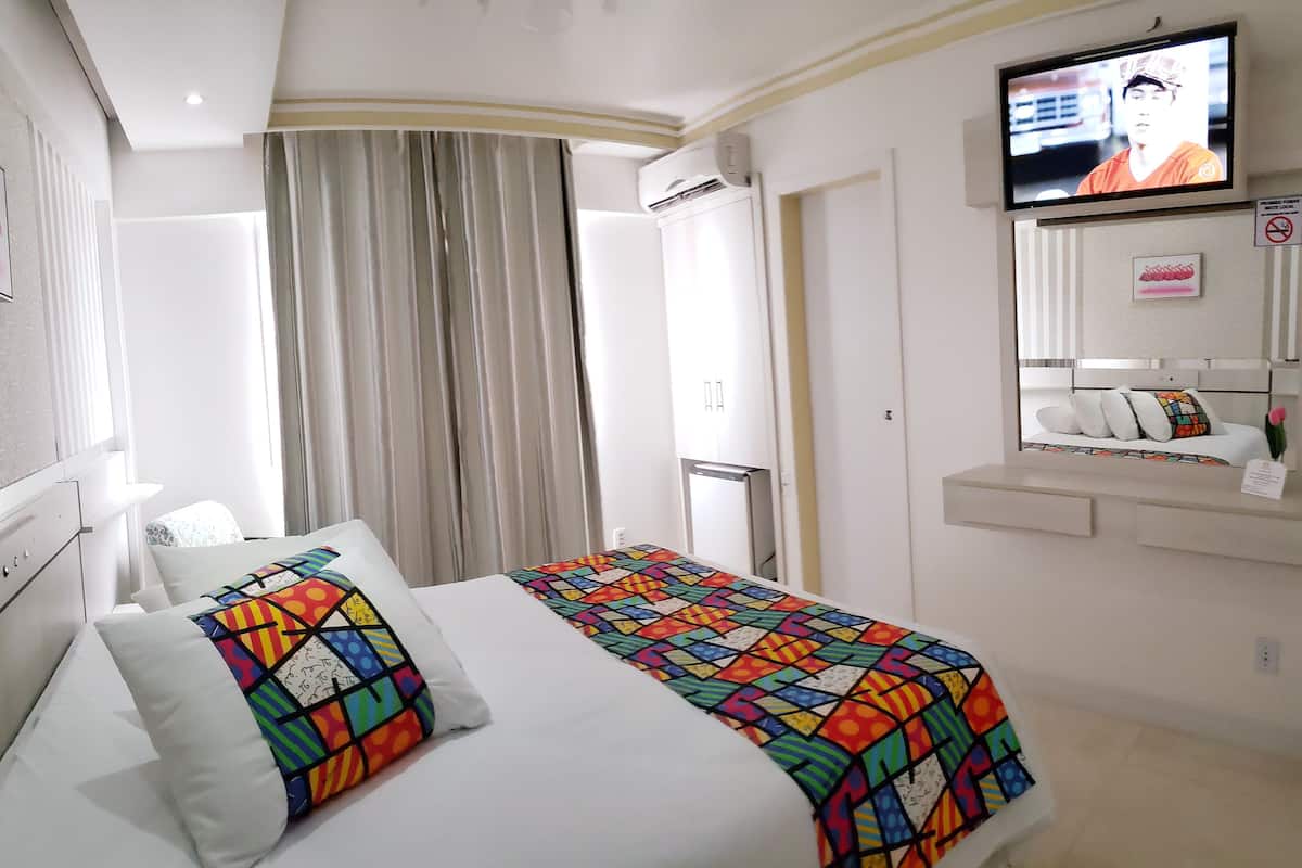 Ein Hotelzimmer mit Bett, Wandfernseher, Fenster mit Vorhängen und einem kleinen Tisch mit einer Vase.