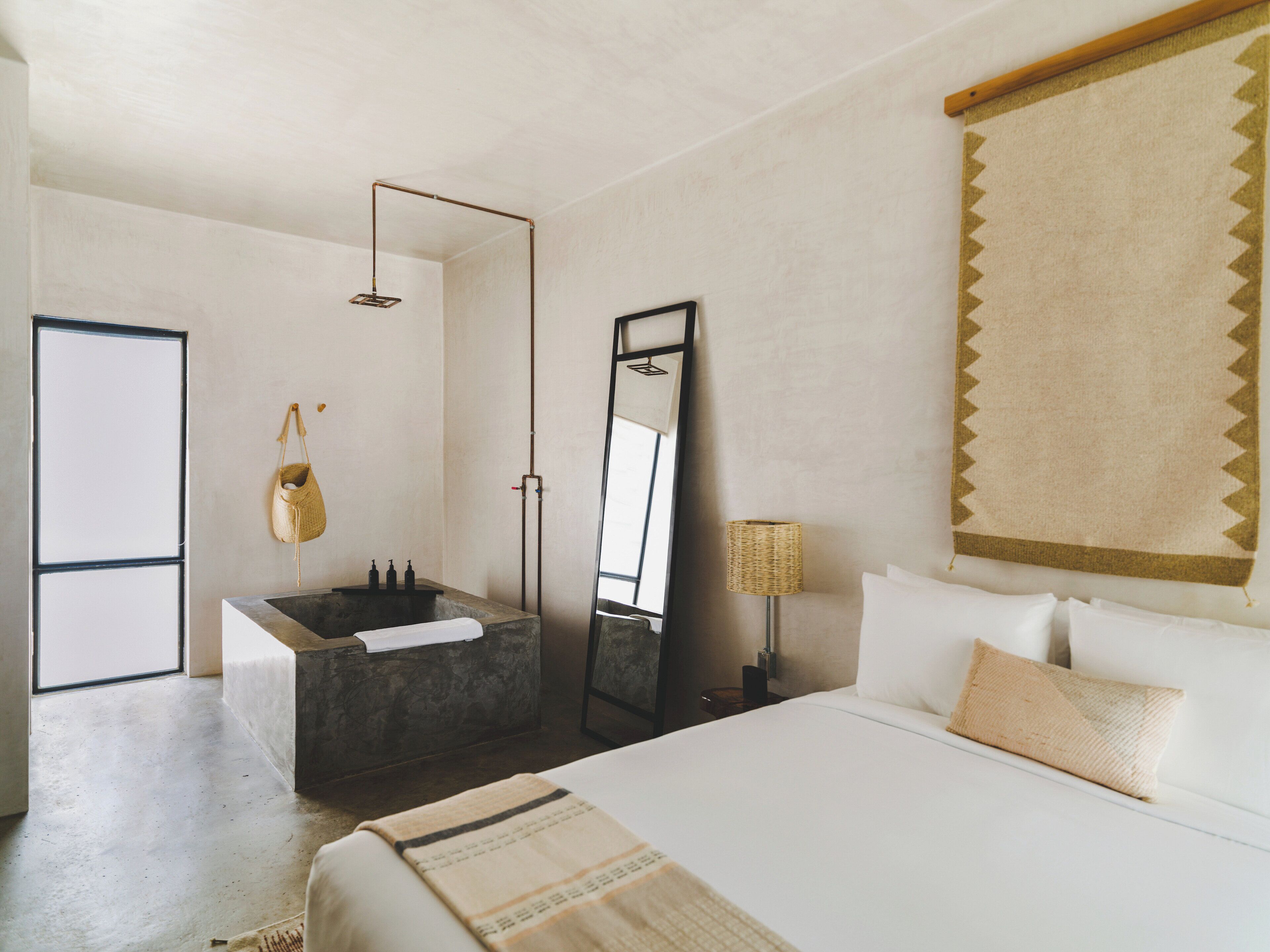Quarto moderno com cama grande, banheira integrada, espelho e cesto suspenso.