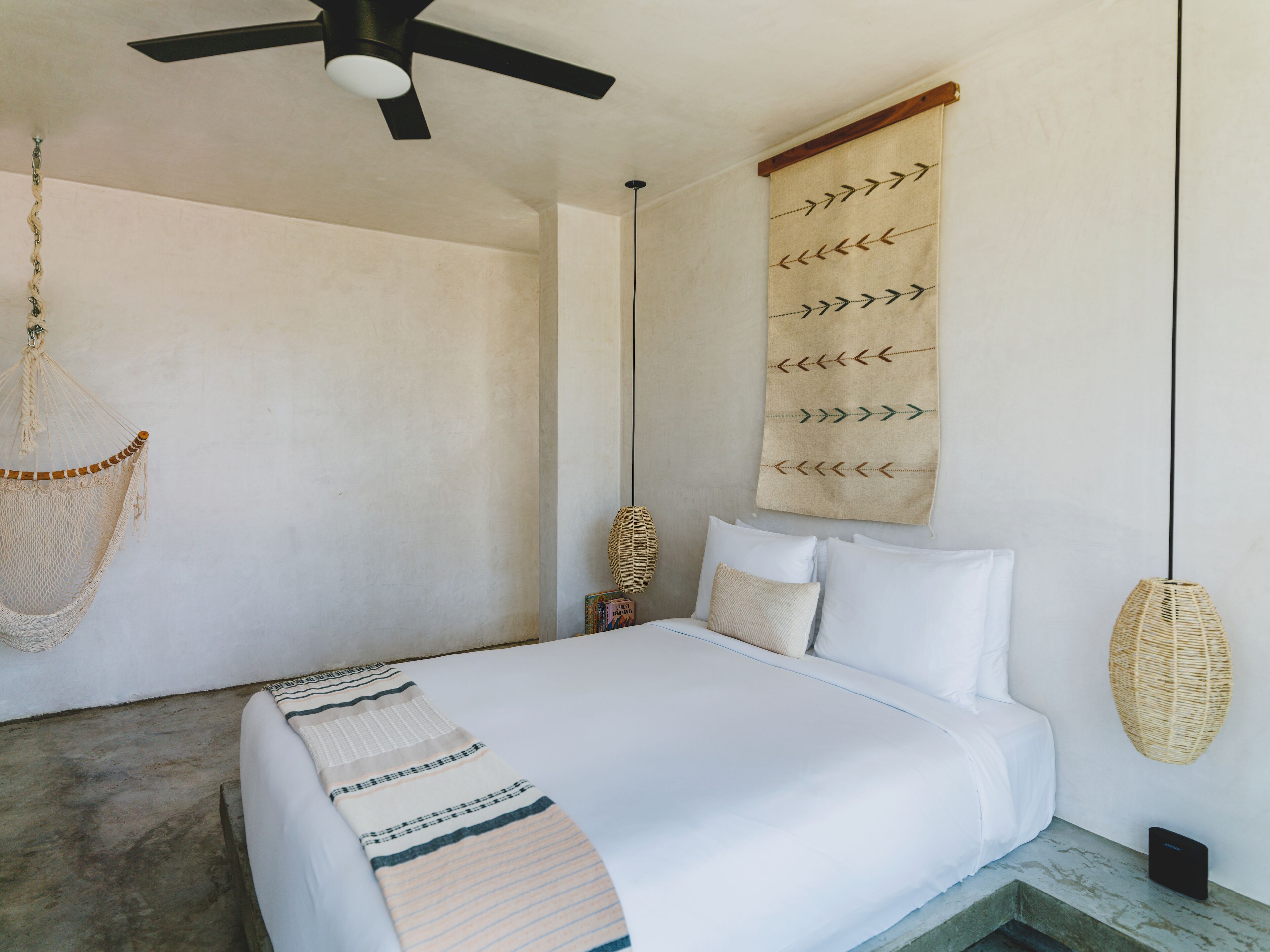 Un dormitorio con cama, una silla colgante, un tapiz de pared tejido y un ventilador de techo.