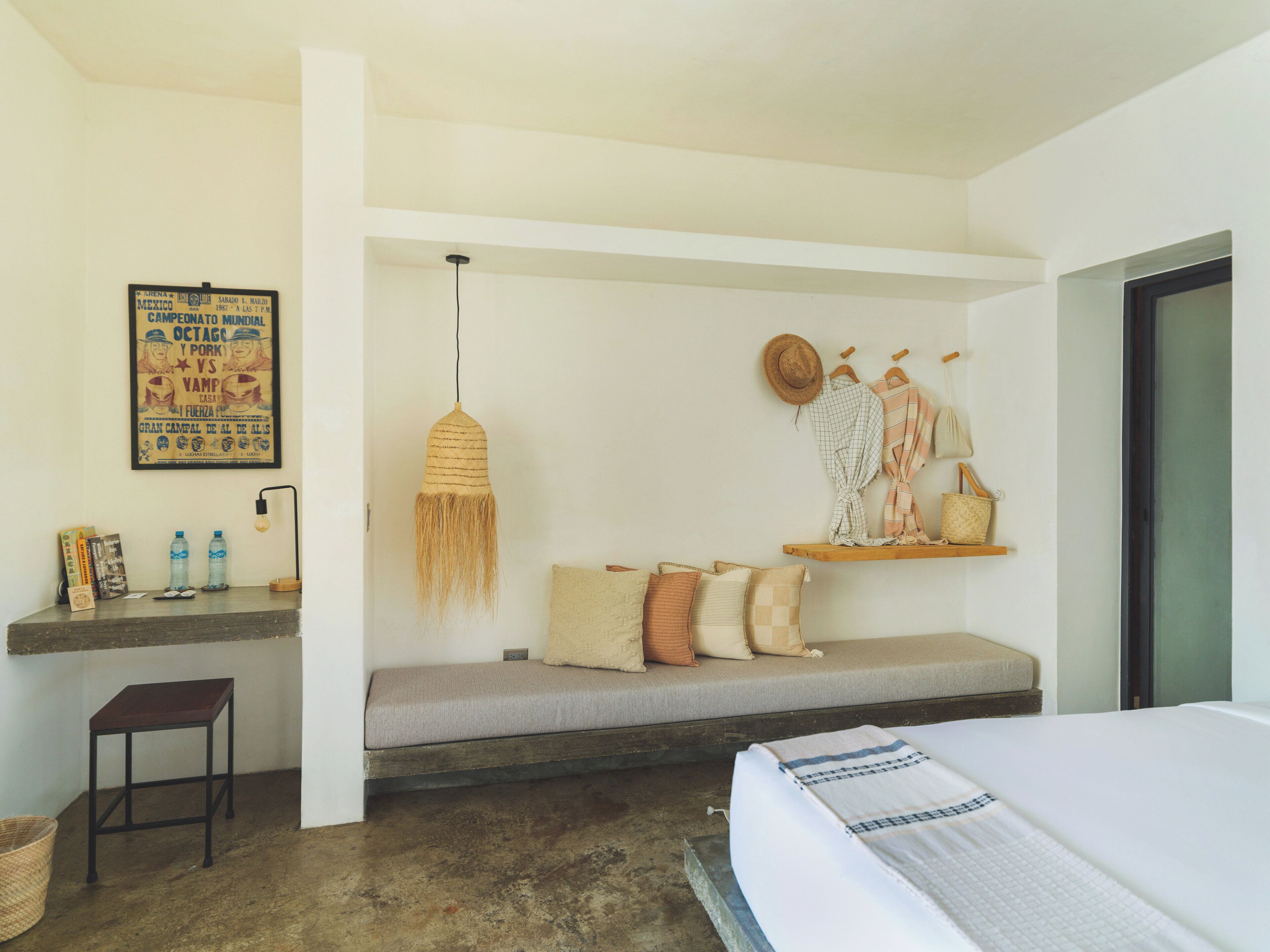 Una habitación de hotel con cama, un banco, una lámpara colgante, un estante con sombreros y un cartel en la pared.