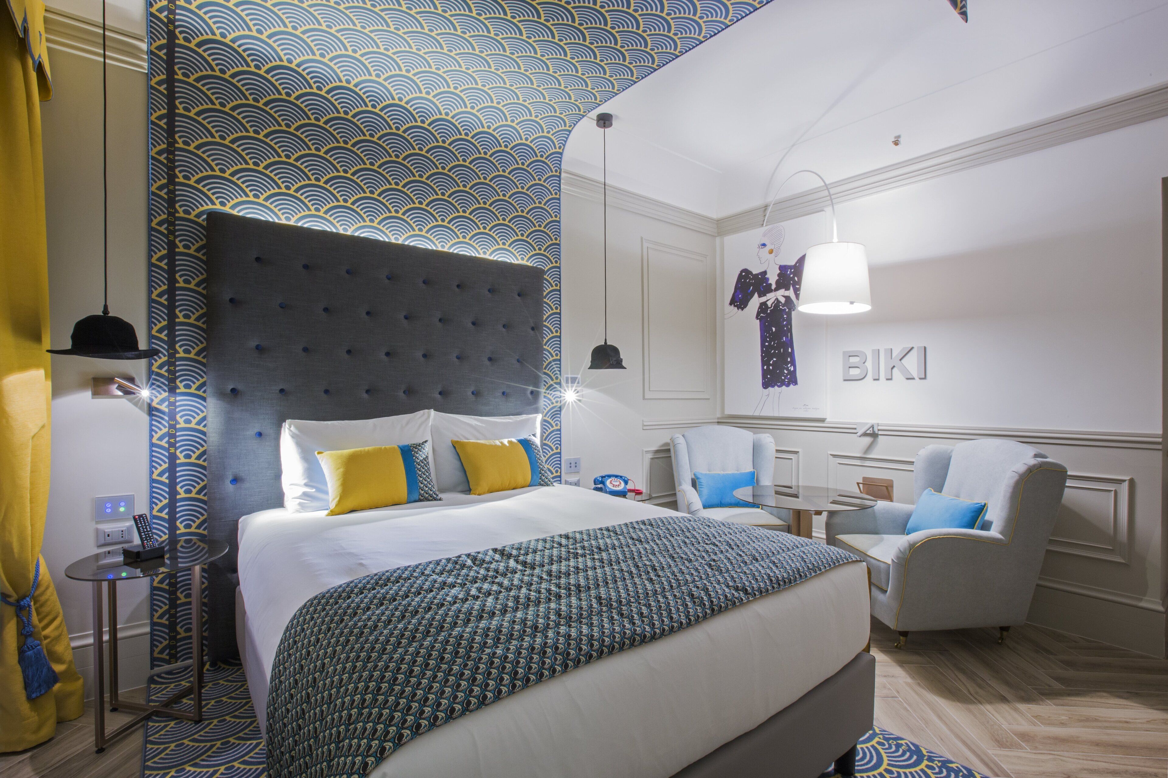 Une chambre d’hôtel avec un grand lit, une tête de lit capitonnée, une petite table avec une lampe, une chaise et un mur orné d’un motif et d’une enseigne sur laquelle on peut lire « BIKI ».