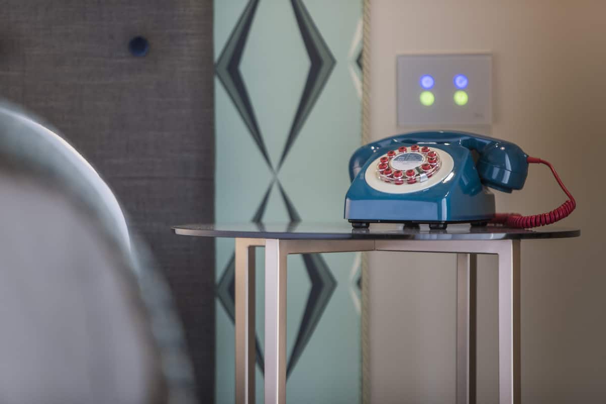 Een blauwe draaischijf telefoon op een klein tafeltje in een kamer met een geometrisch patroon op de muur en een zichtbare lichtschakelaar.