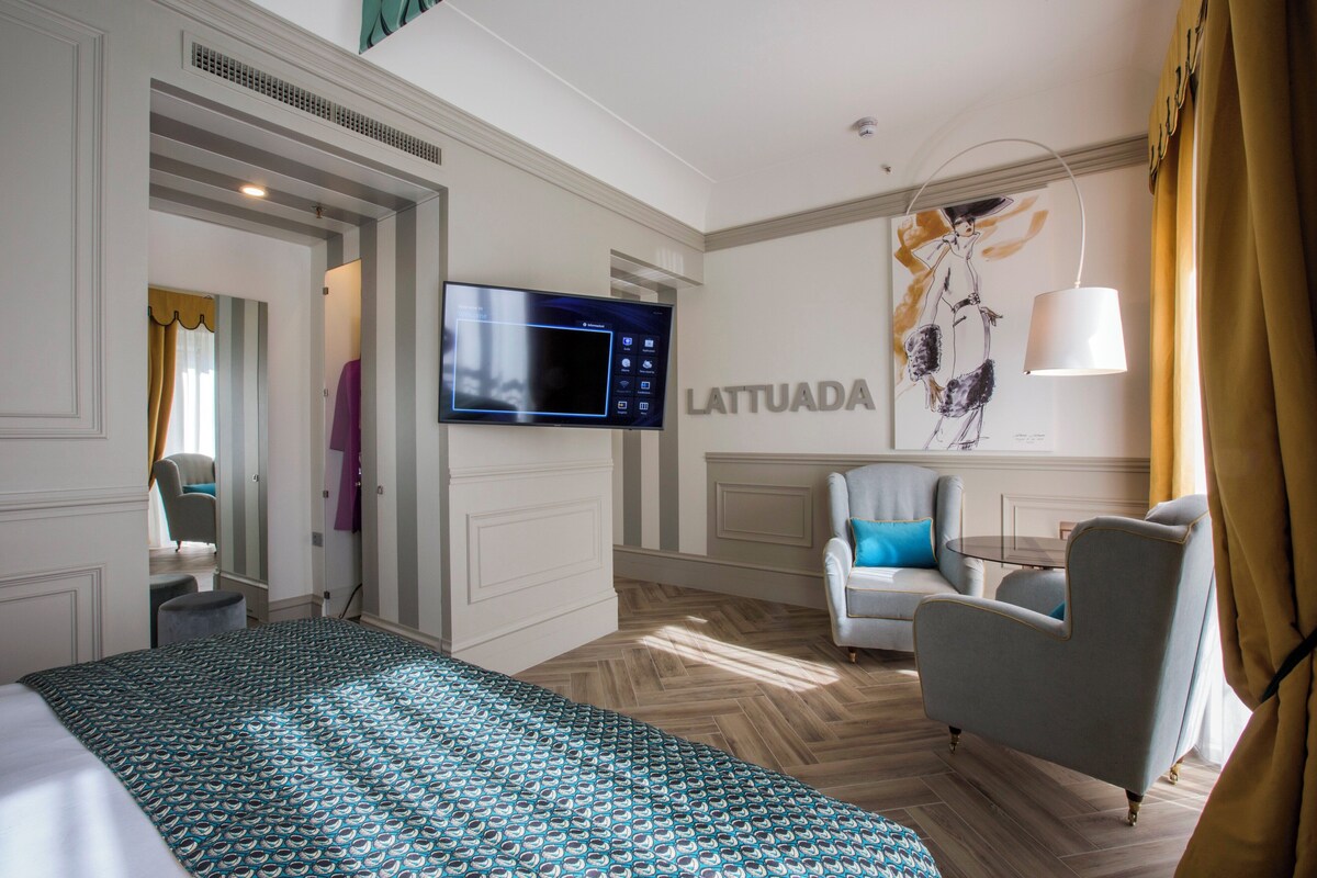 A hotel room with a bed, a seating area with a chair and a table, a flat-screen TV mounted on the wall, and a large artwork on the wall.