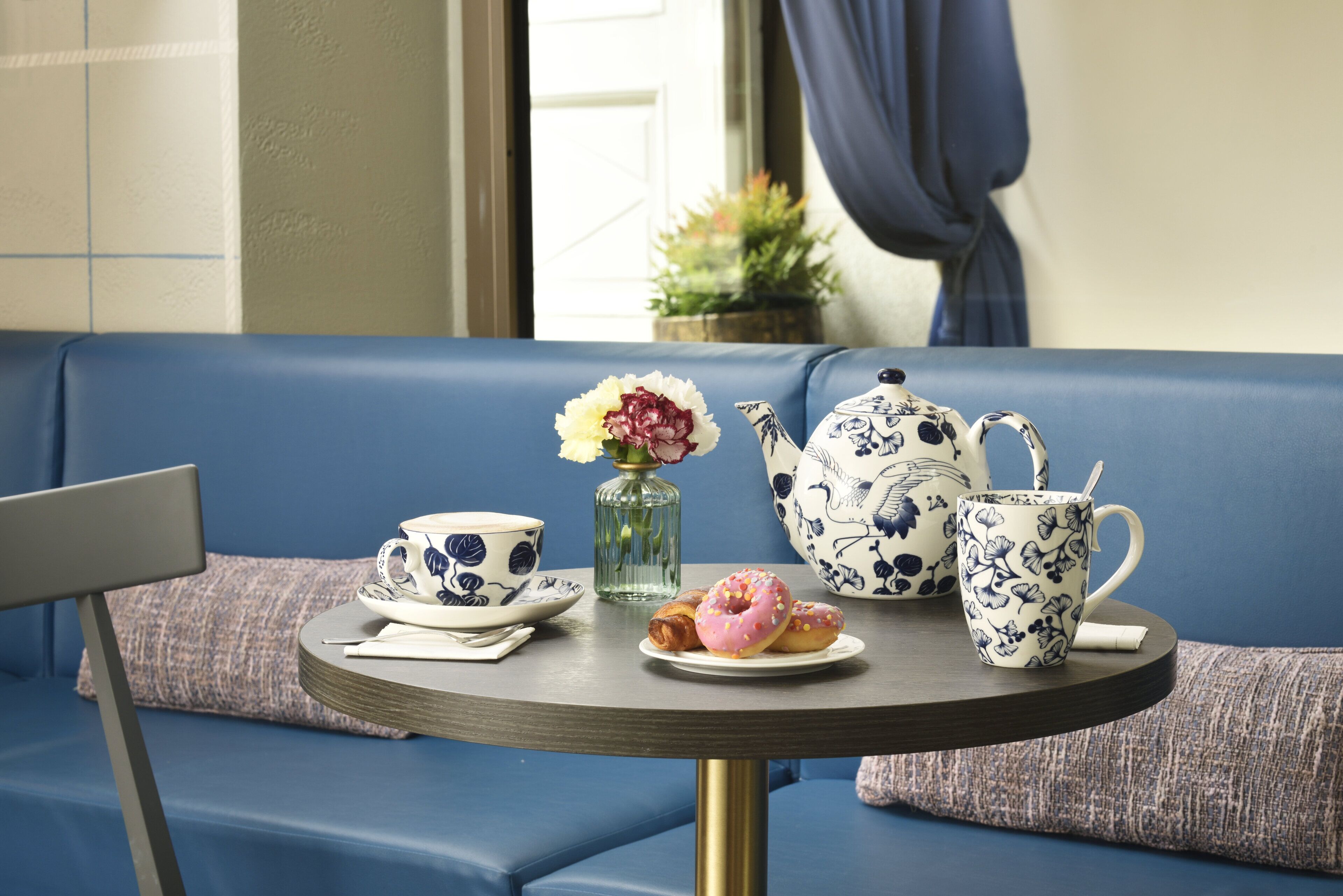 A round table with a teapot, cups, a vase with flowers, and a plate with pastries.