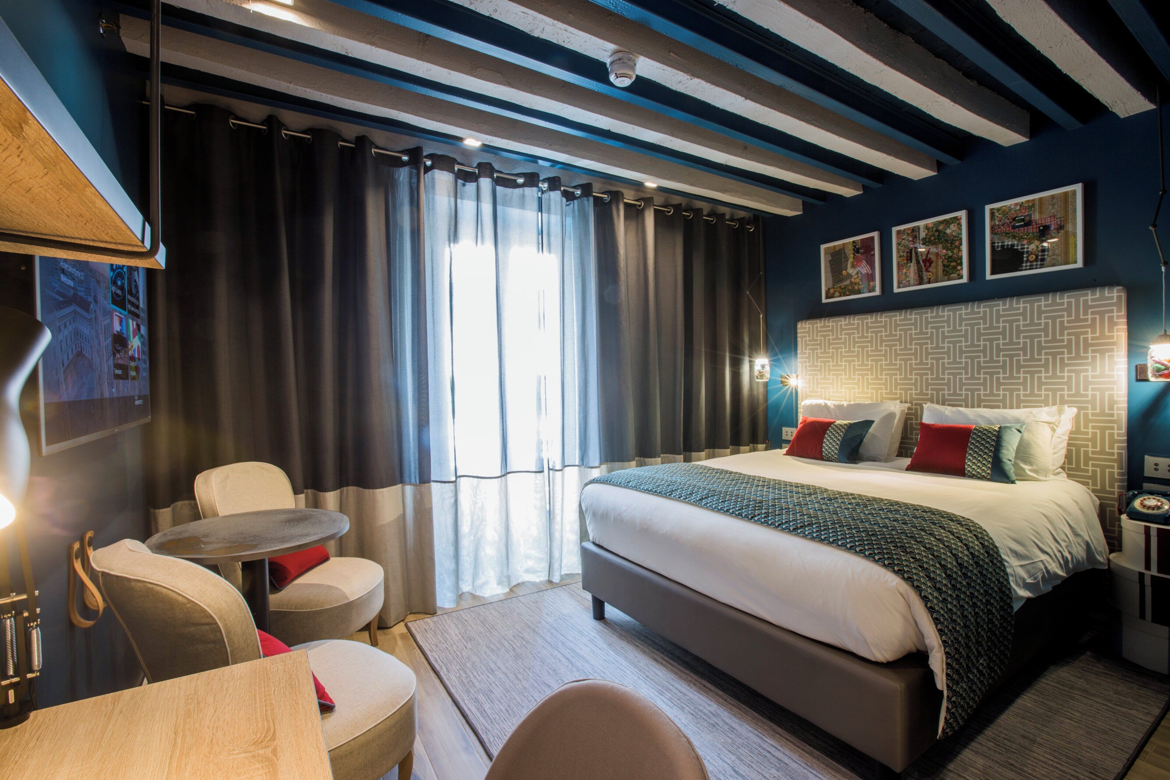 Quarto de hotel com cama, escrivaninha, cadeira, mesa pequena e janela com cortinas.