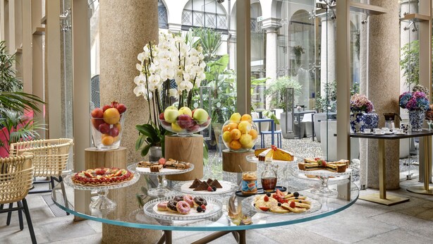 A glass-topped buffet table with various pastries, fruits, and desserts in a spacious indoor area with columns and large windows. - Hotel Indigo Milan - Corso Monforte by IHG (Milan)