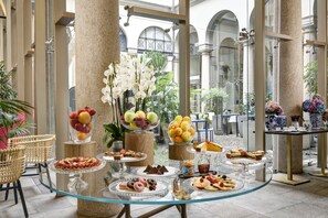A glass-topped buffet table with various pastries, fruits, and desserts in a spacious indoor area with columns and large windows. - Hotel Indigo Milan - Corso Monforte by IHG (Milan)