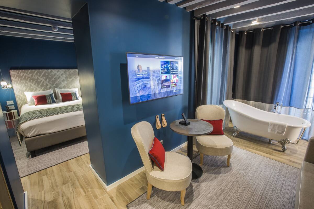 Een moderne hotelkamer met een bed, een zithoek met een tafel, een badkuip en een wandtelevisie waarop een stadsgezicht te zien is.