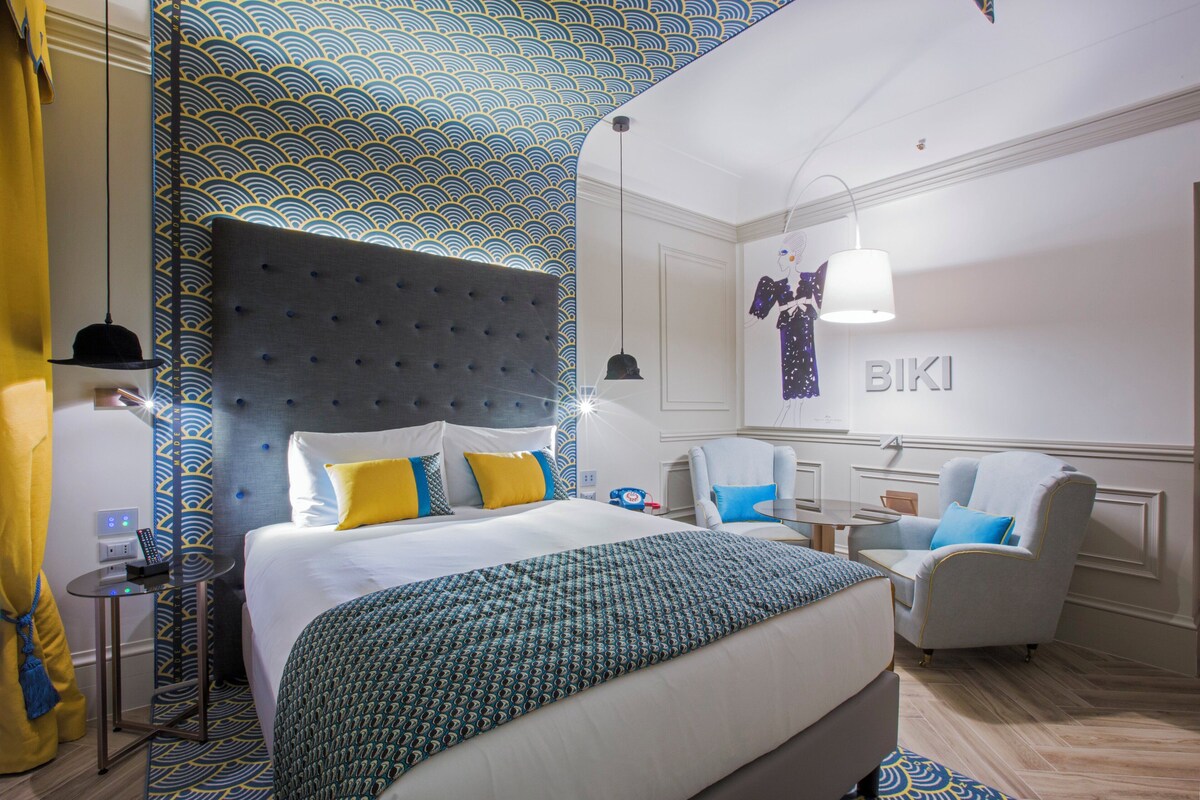 Een hotelkamer met een groot bed, hoofdbord, nachtkastje, stoel, bureau en spiegel.