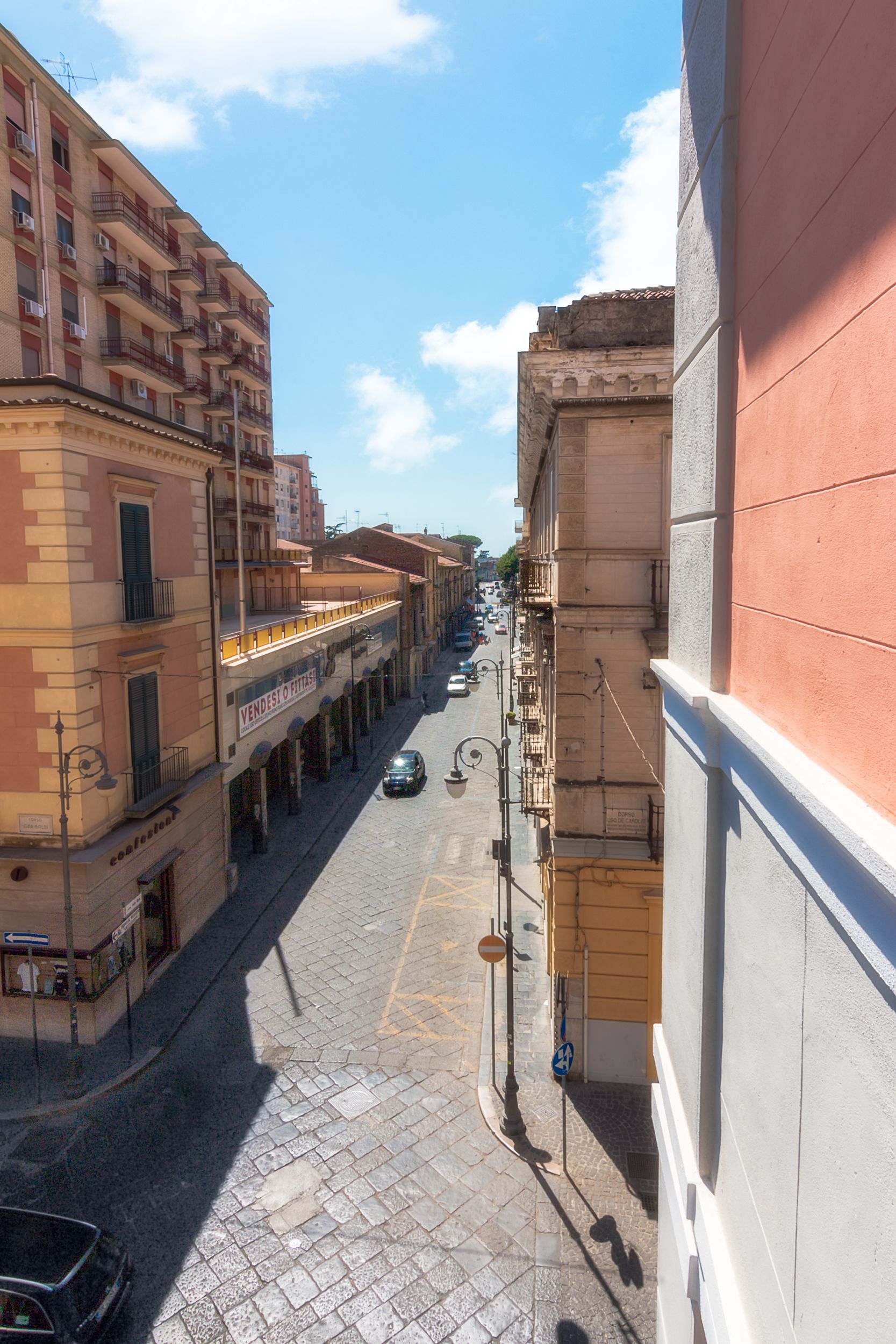 A narrow street lined with multi-story buildings, featuring balconies and a cobblestone pavement.