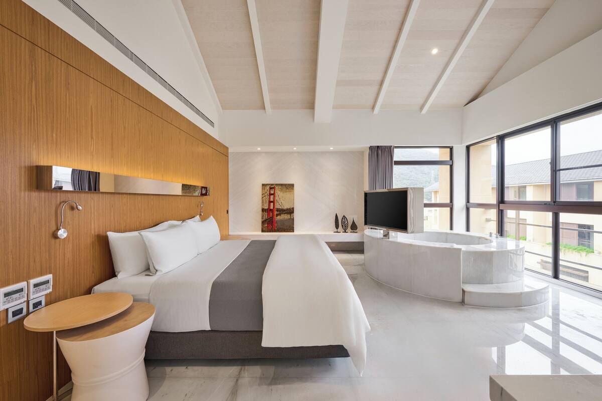 A modern hotel room with a large bed, a curved bathtub, a flat-screen TV, and a large window offering a view of the outside.