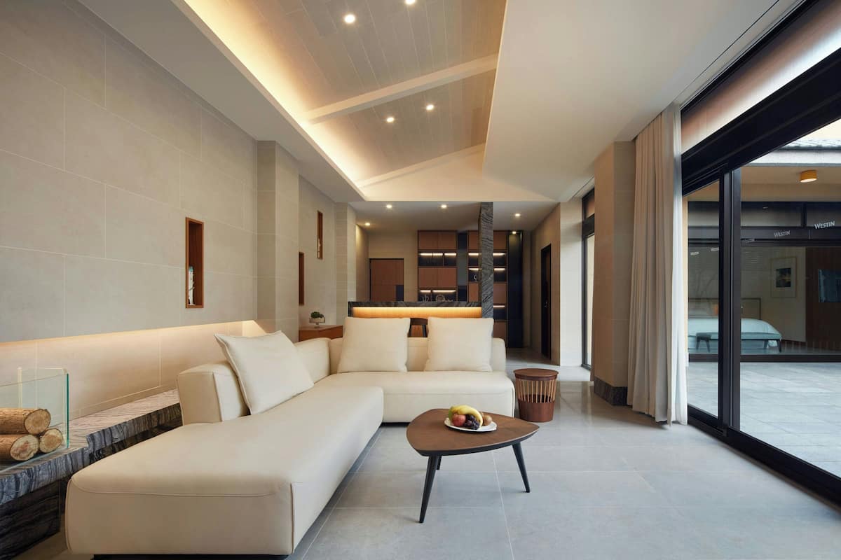 A modern living room with a sectional sofa, a coffee table with fruit, and a view of the street through large windows.