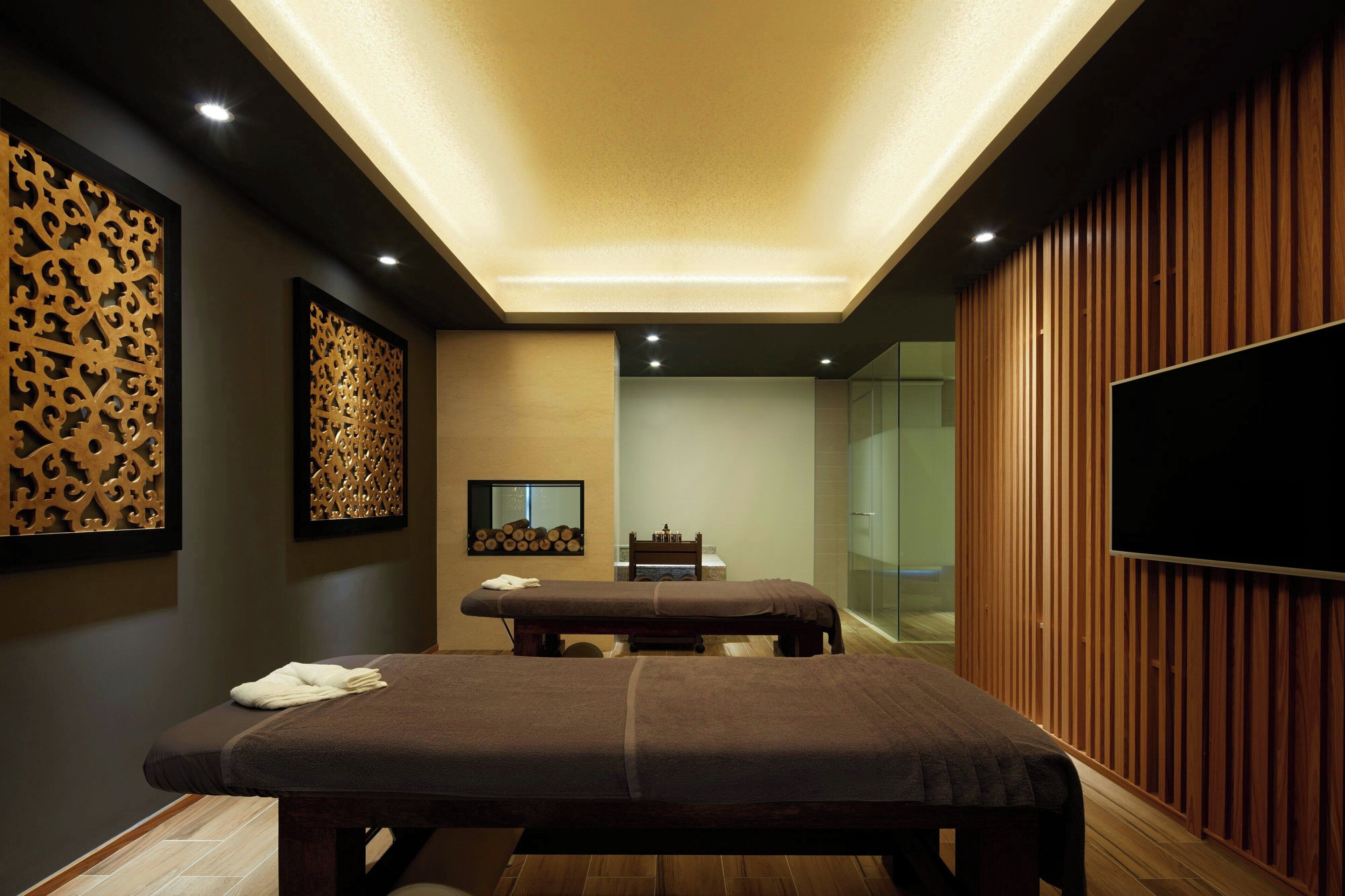 A spa room with two massage tables, wooden paneling, a flat-screen TV, and a wall-mounted mirror.