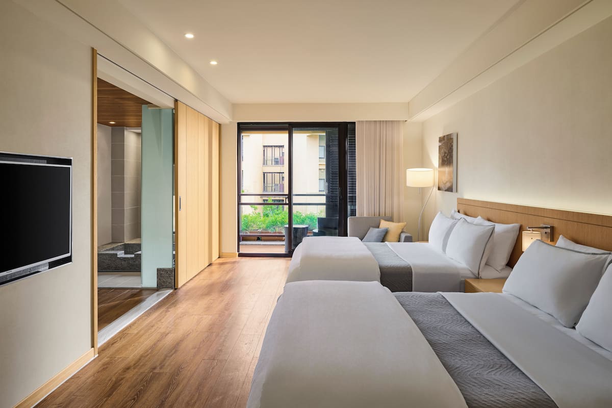 A hotel room with two beds, a TV, a sliding door to a balcony, and a view of the outside.