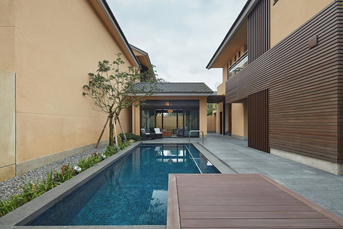 A modern outdoor pool area with a wooden deck, seating, and a covered patio.