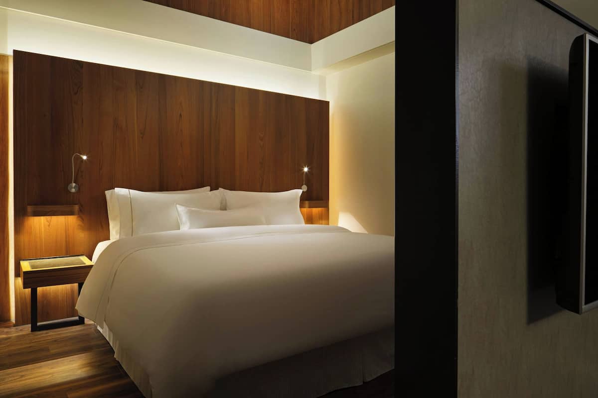 A modern hotel room with a wooden headboard, a large bed with white linens, bedside tables with lamps, and a flat-screen TV mounted on the wall.