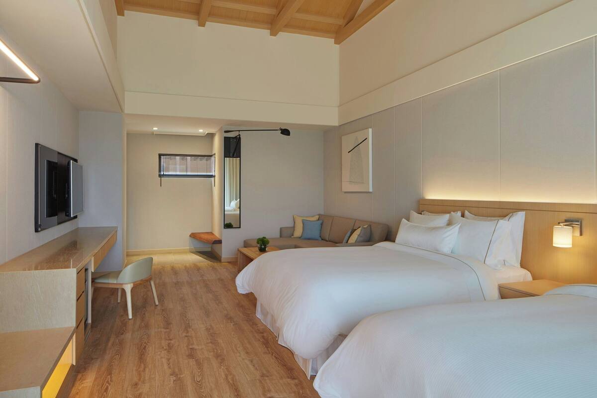 A modern hotel room with two large beds, a desk, a chair, a television, and a small table with a plant.