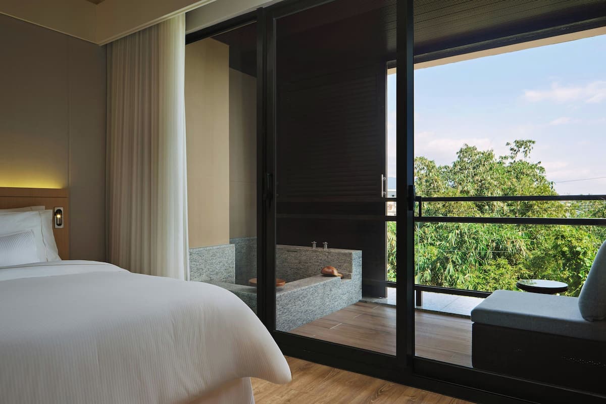 A modern hotel room with a large bed, a bathroom with a bathtub, and a balcony with a view of greenery.