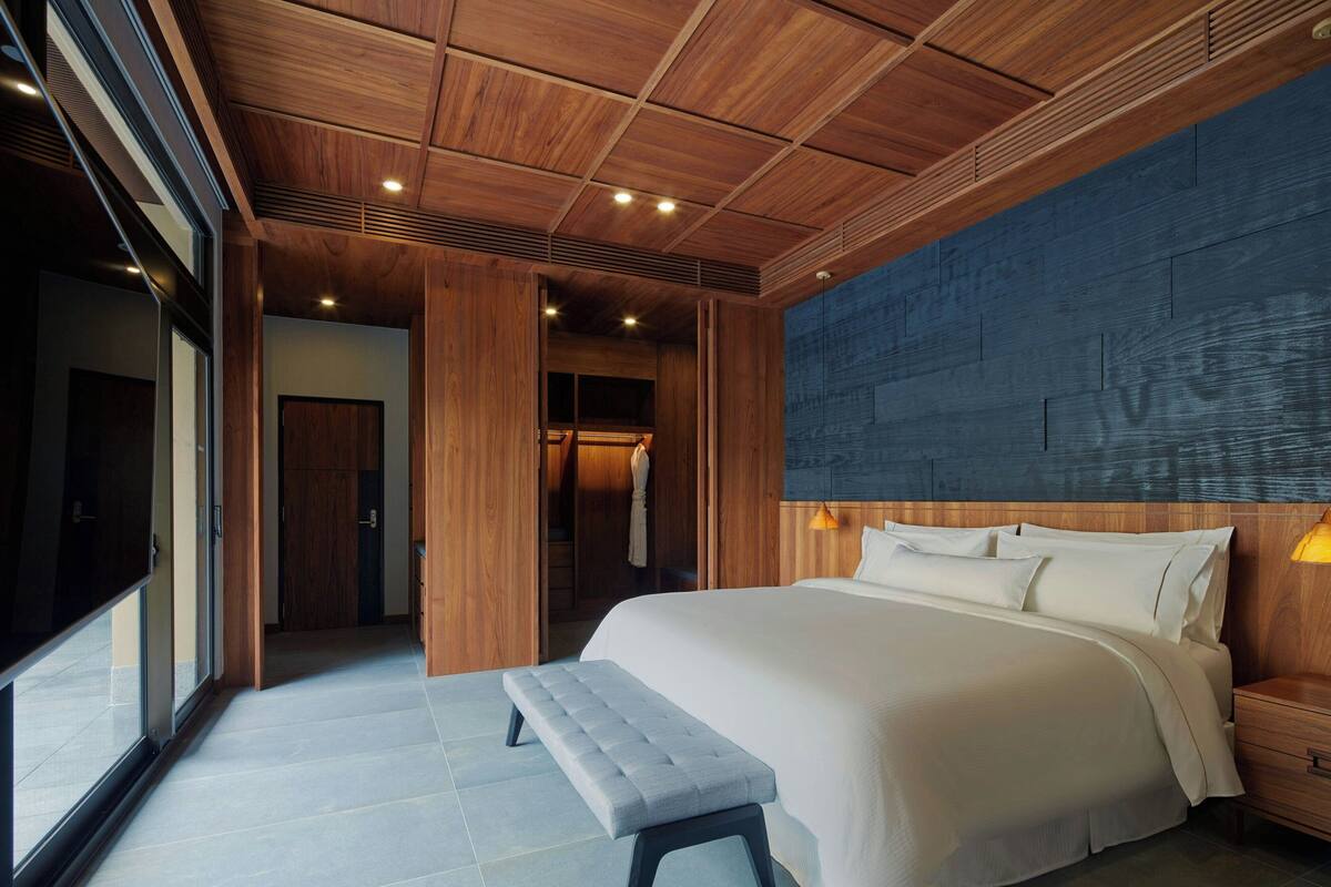A modern bedroom with a large bed, wooden ceiling, and a view through a glass door.