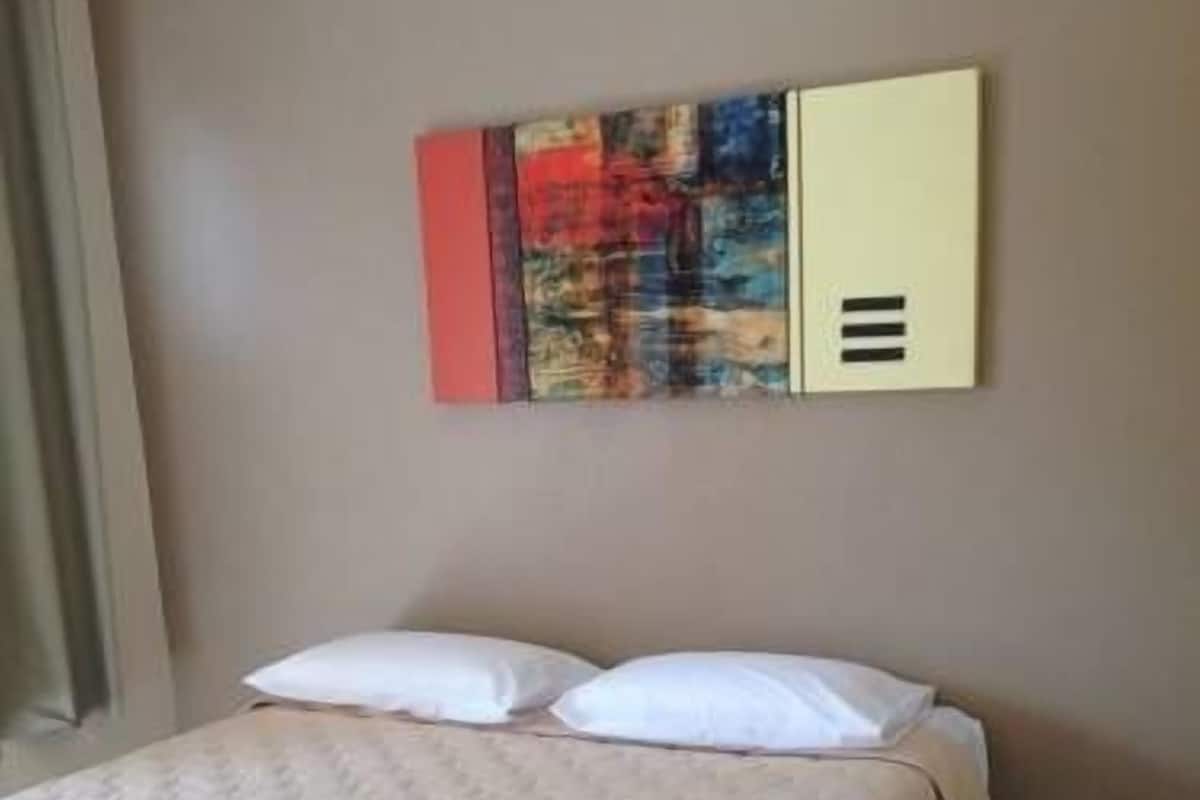 Un lit avec une couette et des oreillers blancs, un mur beige et, au-dessus, un tableau abstrait aux couleurs vives.