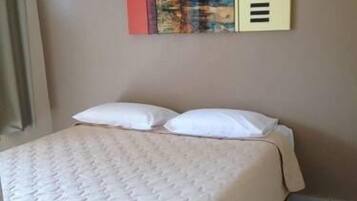 Uma cama com colcha e travesseiros brancos, uma parede bege e uma pintura abstrata colorida em cima.