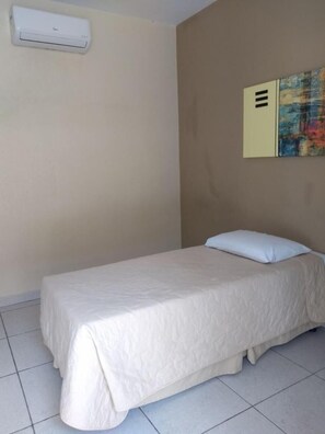 A single bed with a white bedspread in a room with a beige wall, a white air conditioning unit, and a colorful abstract painting on the wall. - Hotel Portal do Eden (Sorocaba)