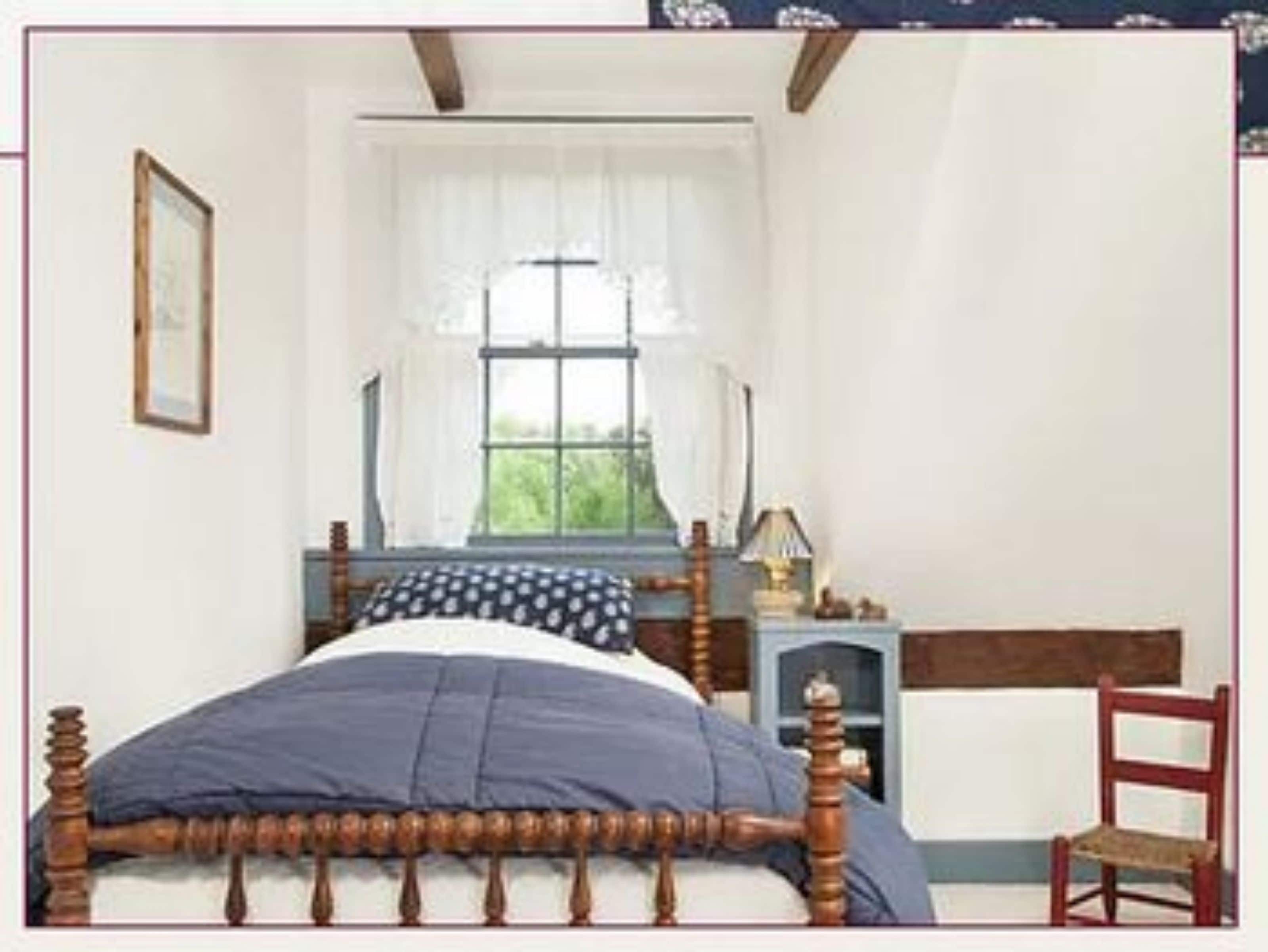 A bedroom with a wooden bed, a window with curtains, a bedside table with a lamp, and a chair.