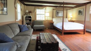 Un dormitorio con una cama con dosel, un sofá, un sillón pequeño, una mesita de noche con lámpara, una estantería y un ventanal con cortinas.
