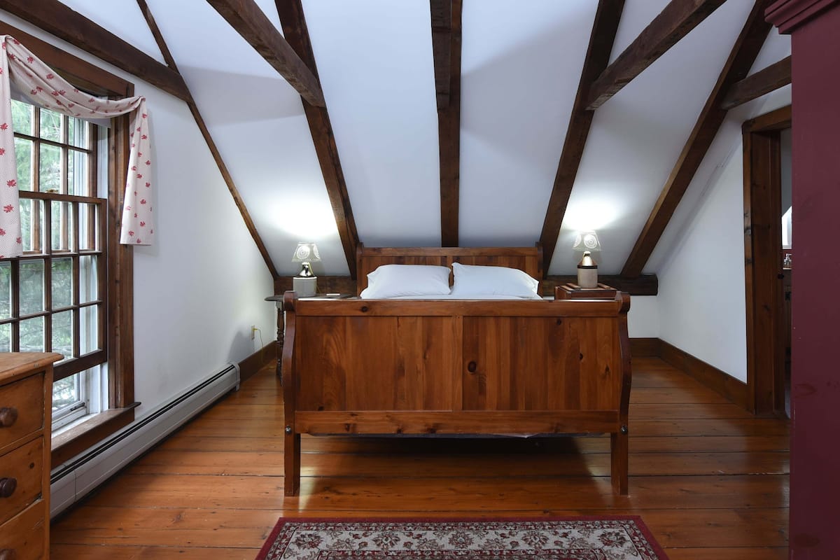 A bedroom with a wooden bed, a nightstand with a lamp, a window with curtains, and a radiator.