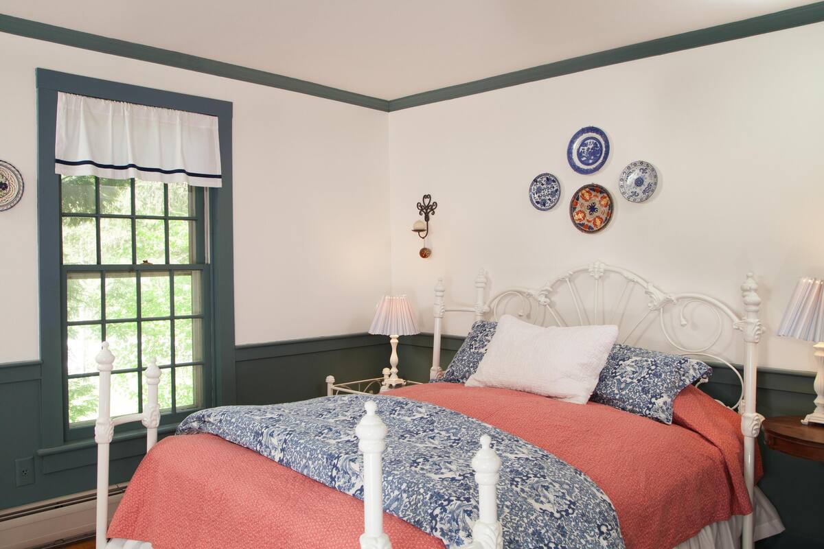 A bedroom with a white four-poster bed, a window with a white valance, and a wall with decorative plates.