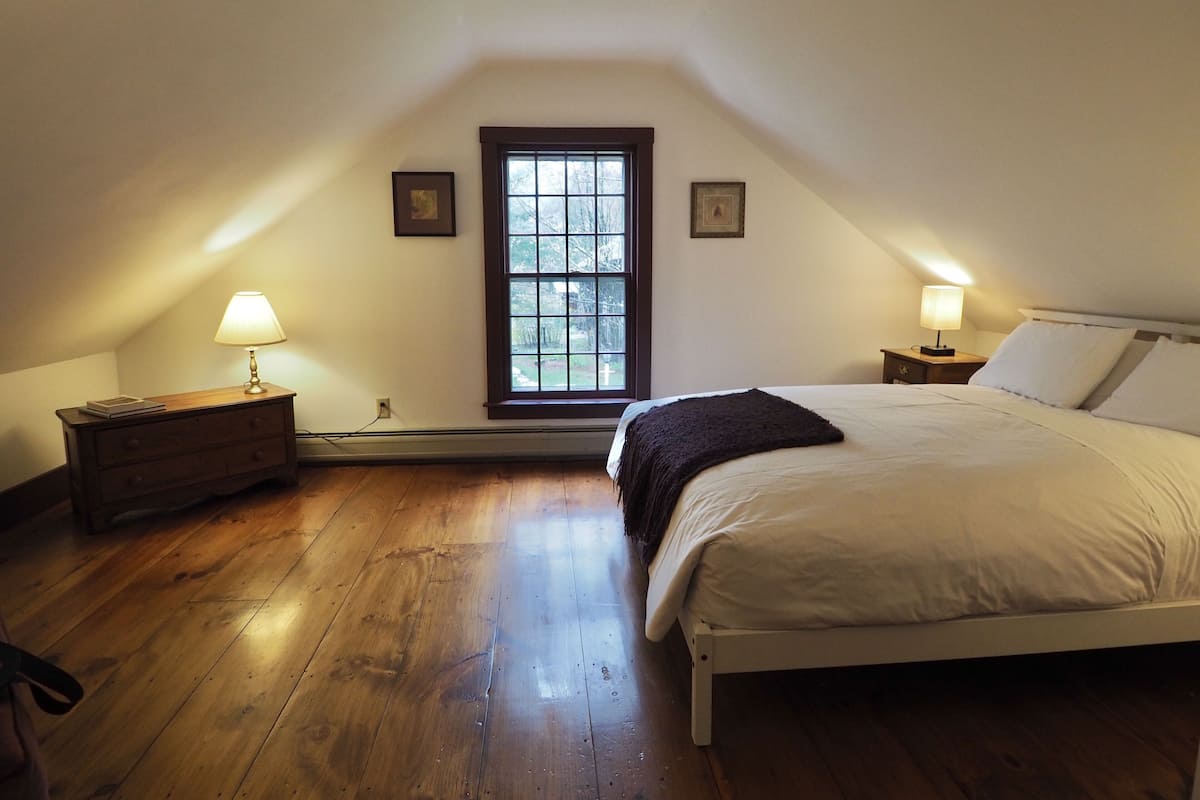 A bedroom with a large window, a bed, a nightstand with a lamp, and a dresser.