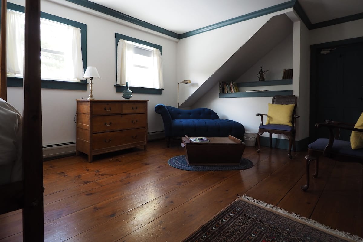 A cozy living room with a wooden dresser, a blue sofa, a small coffee table, and a chair with a yellow cushion. There is a window with curtains, a lamp, and a shelf with books and a decorative item.