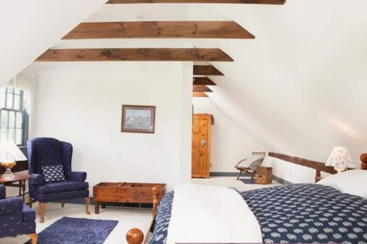 A room with a sloped ceiling, a bed with patterned bedding, a blue armchair, a wooden chest of drawers, and a framed picture on the wall.