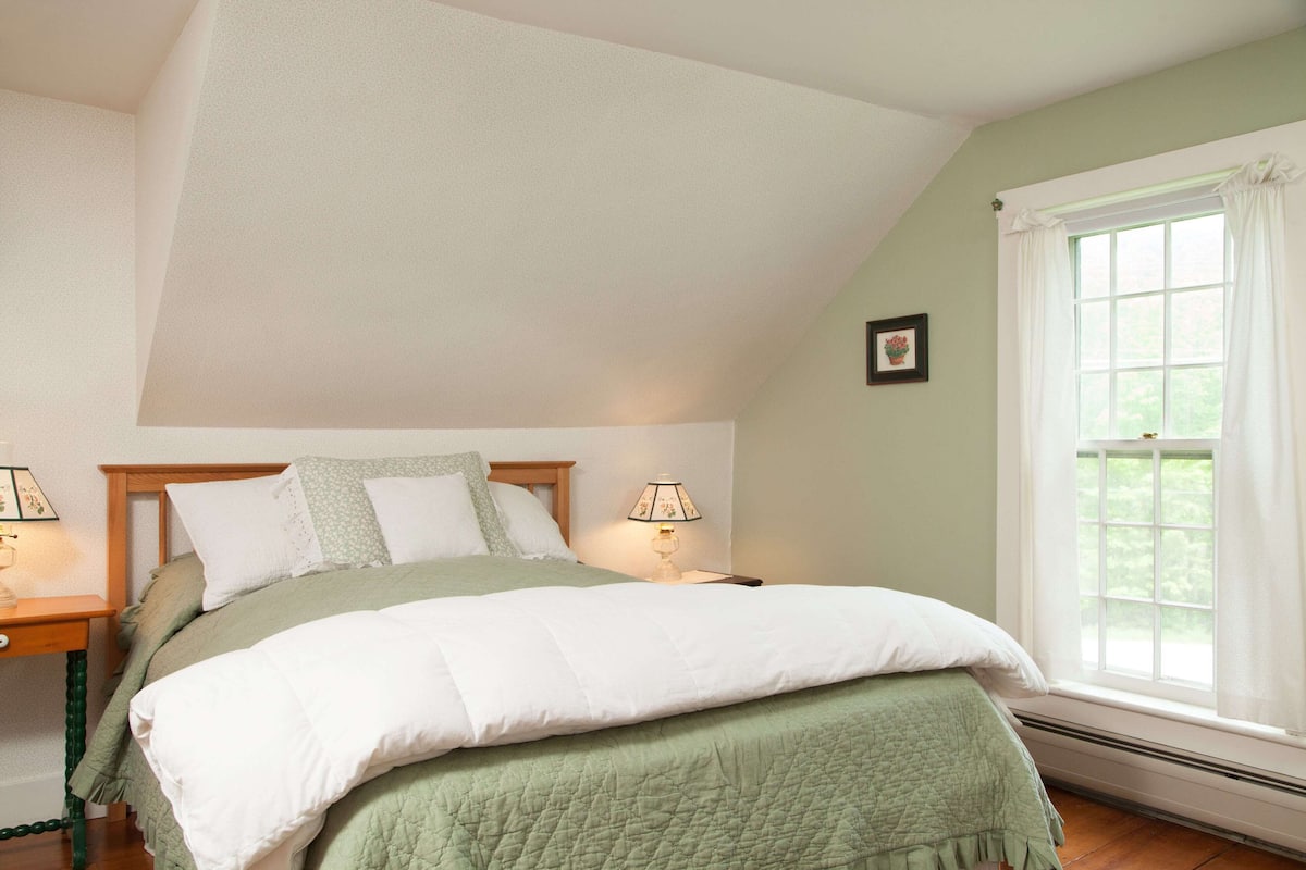 A bedroom with a sloped ceiling, a bed with green bedding, two bedside lamps, a window with white curtains, and a framed picture on the wall.
