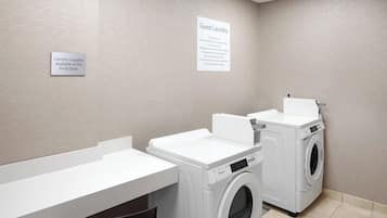 A hotel laundry room with two front-loading washing machines, a clock, and signs indicating guest laundry services and available supplies at the front desk.