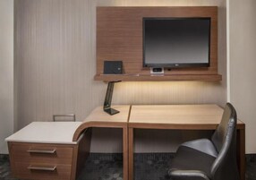 Habitación de hotel con un escritorio de madera, un televisor de pantalla plana, una lámpara, una silla y un altavoz pequeño.