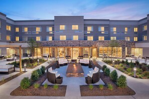 A modern hotel courtyard with a central fire pit surrounded by outdoor seating, stone walls, and a well-lit entrance. - Courtyard by Marriott Wayne Fairfield (Wayne)