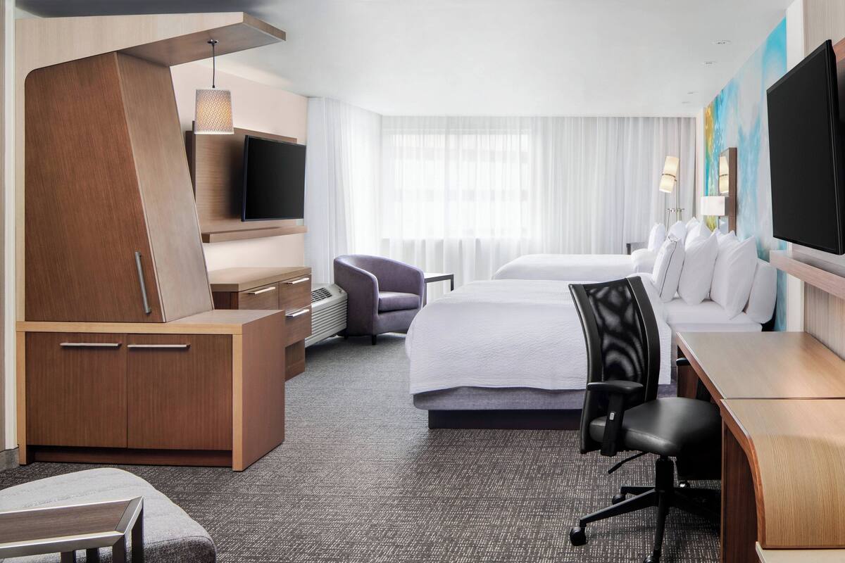 A hotel room with a bed, two chairs, a desk, a TV, and a wardrobe.